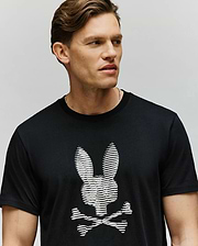 A man with short brown hair wears the Psycho Bunny MENS GRANT BUNNY GRAPHIC TEE - MT0200081, a soft Pima cotton black tee with an HD-printed white bunny head and crossed bones. He faces sideways against a plain background.