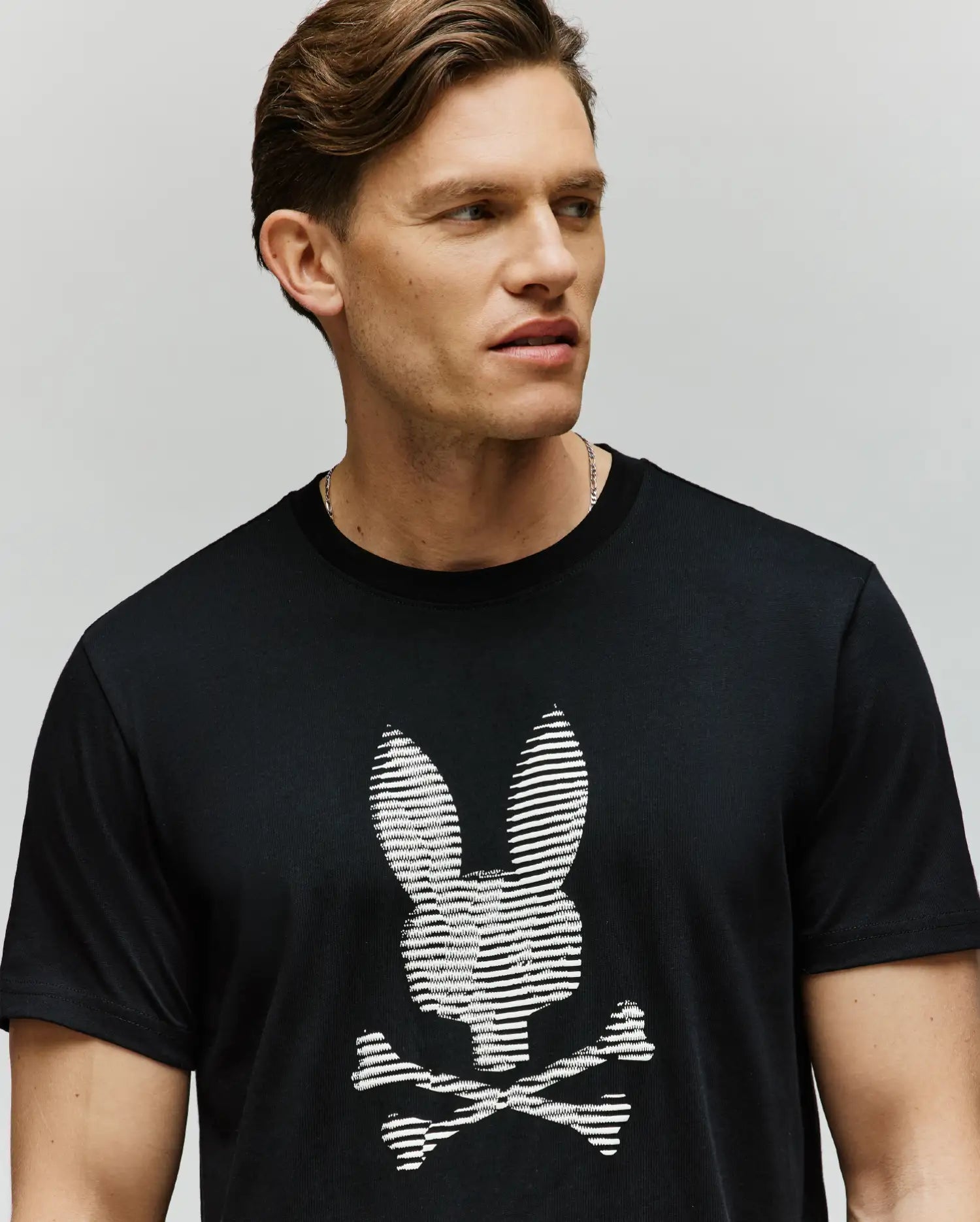 A man with short brown hair wears the Psycho Bunny MENS GRANT BUNNY GRAPHIC TEE - MT0200081, a soft Pima cotton black tee with an HD-printed white bunny head and crossed bones. He faces sideways against a plain background.