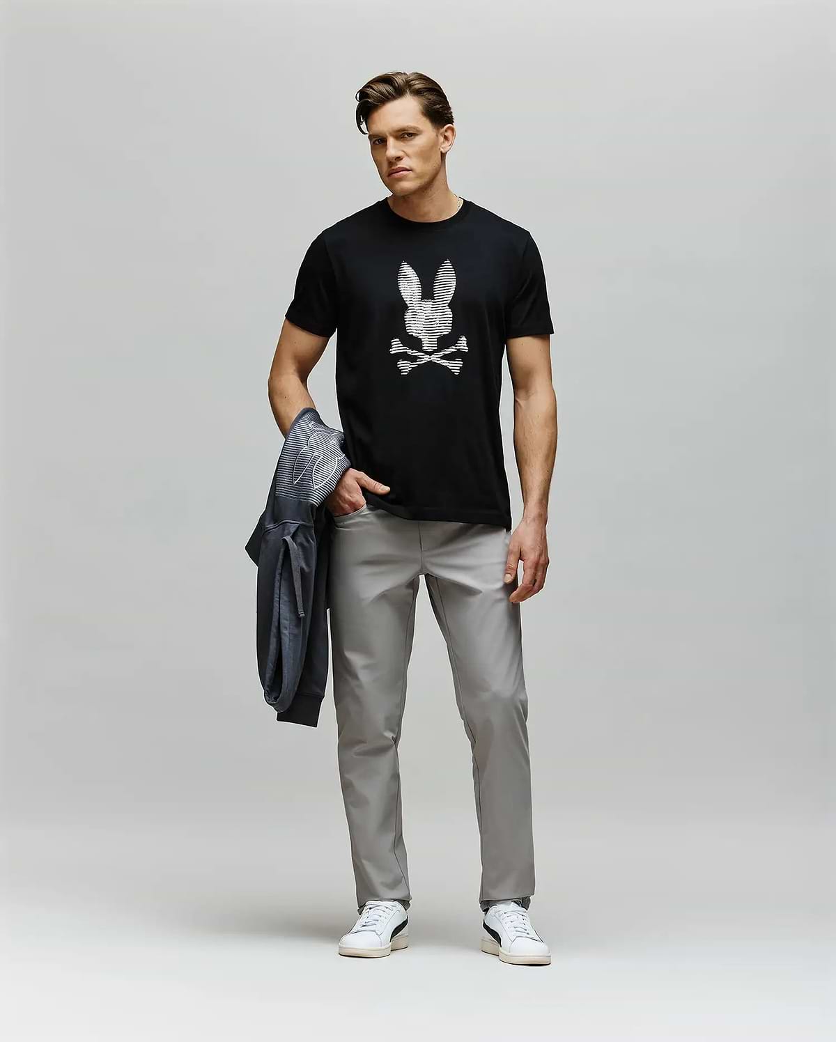 A man stands against a plain background wearing the Psycho Bunny MENS GRANT BUNNY GRAPHIC TEE - MT0200081, featuring a skull and crossbones design, paired with light gray pants, white sneakers, and holding a gray jacket in his left hand.