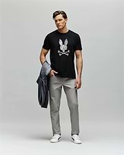 A man stands against a plain background wearing the Psycho Bunny MENS GRANT BUNNY GRAPHIC TEE - MT0200081, featuring a skull and crossbones design, paired with light gray pants, white sneakers, and holding a gray jacket in his left hand.