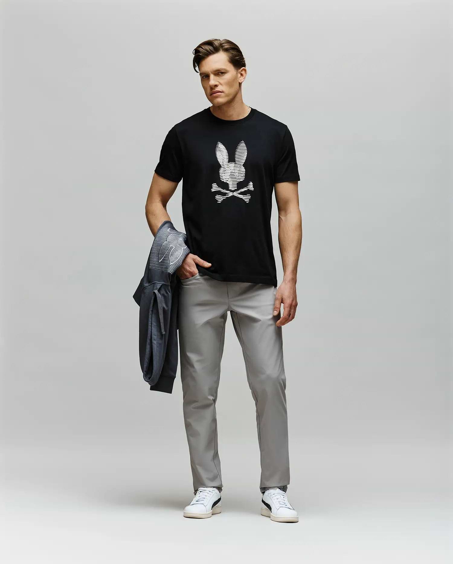 A man stands against a plain background wearing the Psycho Bunny MENS GRANT BUNNY GRAPHIC TEE - MT0200081, featuring a skull and crossbones design, paired with light gray pants, white sneakers, and holding a gray jacket in his left hand.