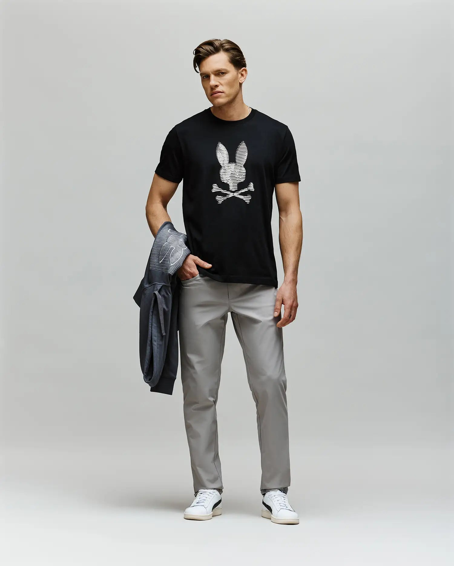 A man stands against a plain background wearing the Psycho Bunny MENS GRANT BUNNY GRAPHIC TEE - MT0200081, featuring a skull and crossbones design, paired with light gray pants, white sneakers, and holding a gray jacket in his left hand.