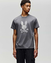A young man with short curly hair stands against a light gray background, wearing the Psycho Bunny MENS GRANT BUNNY GRAPHIC TEE (MT0200081), crafted in Peru from soft Pima cotton. He gazes thoughtfully to the side. (2)