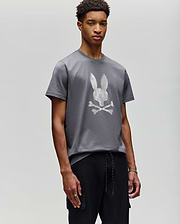 A young man stands against a plain background, wearing Psycho Bunny’s MENS GRANT BUNNY GRAPHIC TEE - MT0200081 in gray with a white pixelated bunny and crossbones graphic, paired with black pants, looking calmly at the camera. (4)