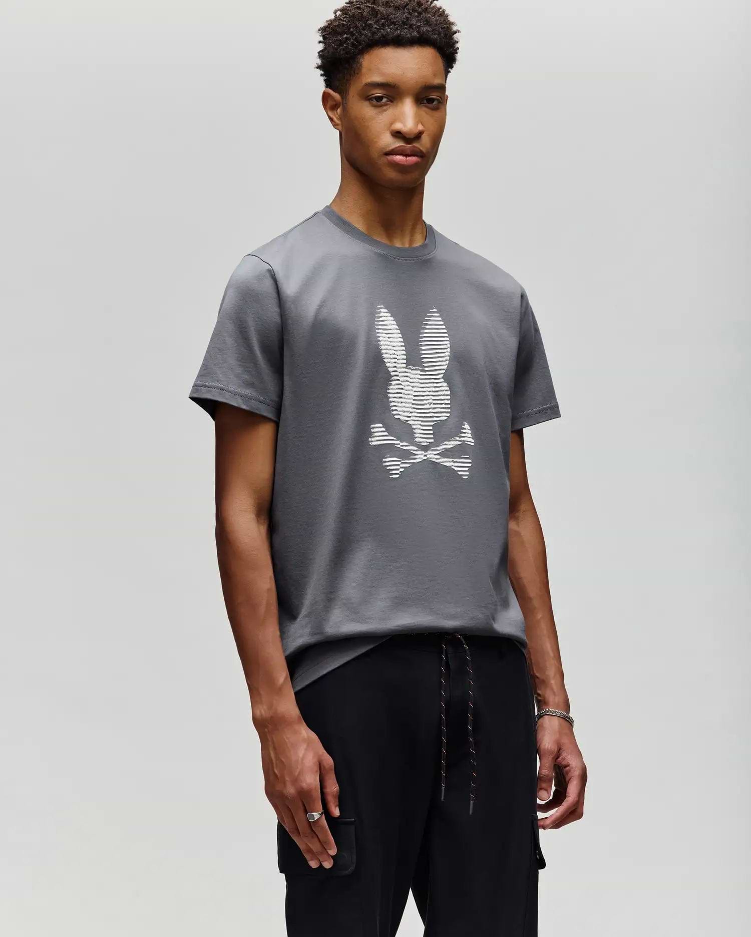 A young man stands against a plain background, wearing Psycho Bunny’s MENS GRANT BUNNY GRAPHIC TEE - MT0200081 in gray with a white pixelated bunny and crossbones graphic, paired with black pants, looking calmly at the camera. (4)