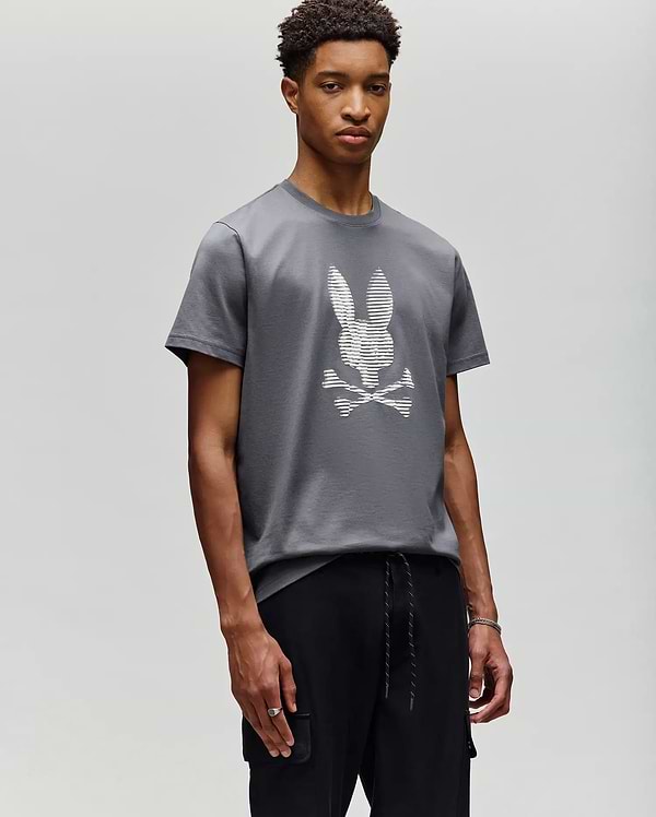 A young man stands against a plain background, wearing Psycho Bunny’s MENS GRANT BUNNY GRAPHIC TEE - MT0200081 in gray with a white pixelated bunny and crossbones graphic, paired with black pants, looking calmly at the camera. (4)