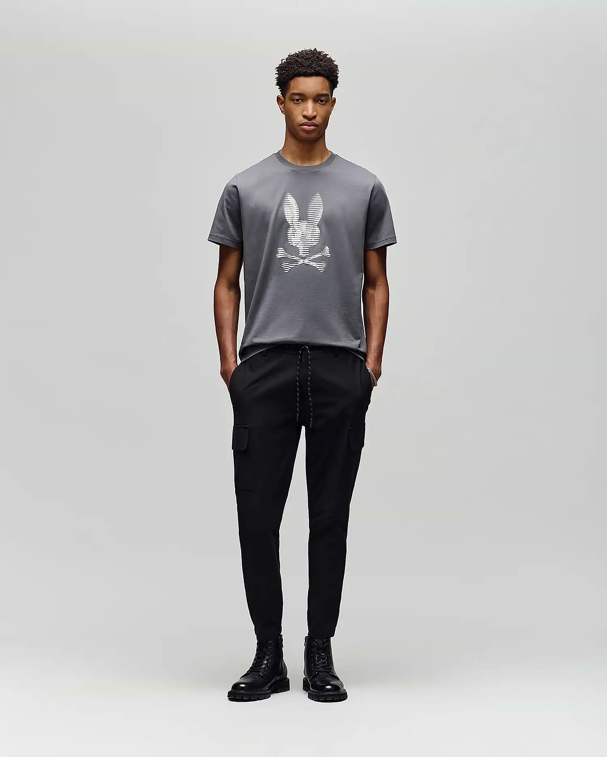 A person poses against a light gray background in the Psycho Bunny MENS GRANT BUNNY GRAPHIC TEE - MT0200081, crafted in Peru from soft Pima cotton, paired with black cargo pants and black lace-up boots. (5)