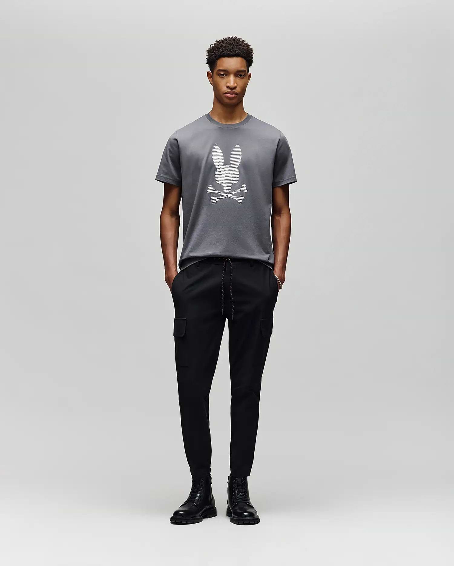A person poses against a light gray background in the Psycho Bunny MENS GRANT BUNNY GRAPHIC TEE - MT0200081, crafted in Peru from soft Pima cotton, paired with black cargo pants and black lace-up boots. (5)