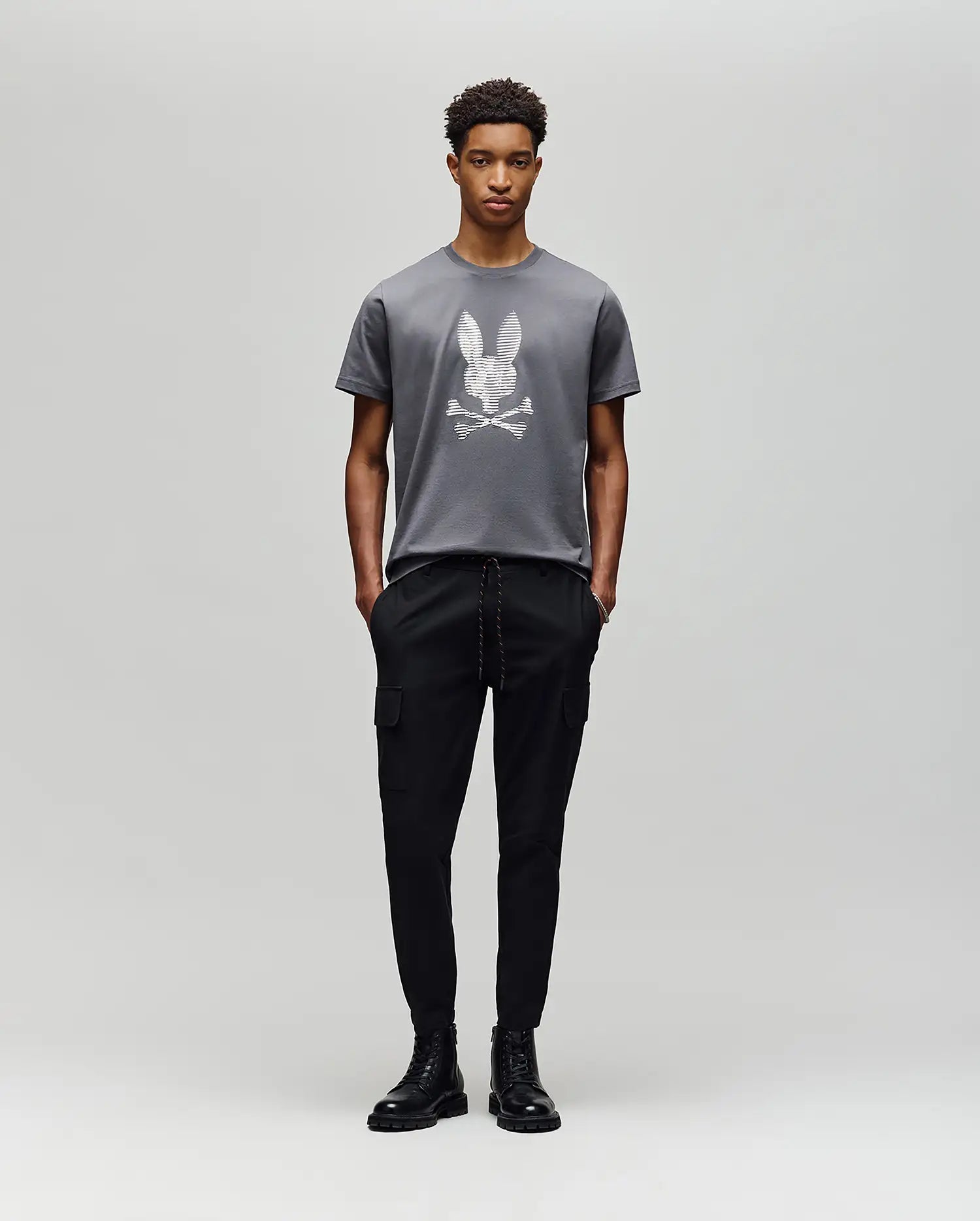A person poses against a light gray background in the Psycho Bunny MENS GRANT BUNNY GRAPHIC TEE - MT0200081, crafted in Peru from soft Pima cotton, paired with black cargo pants and black lace-up boots. (5)