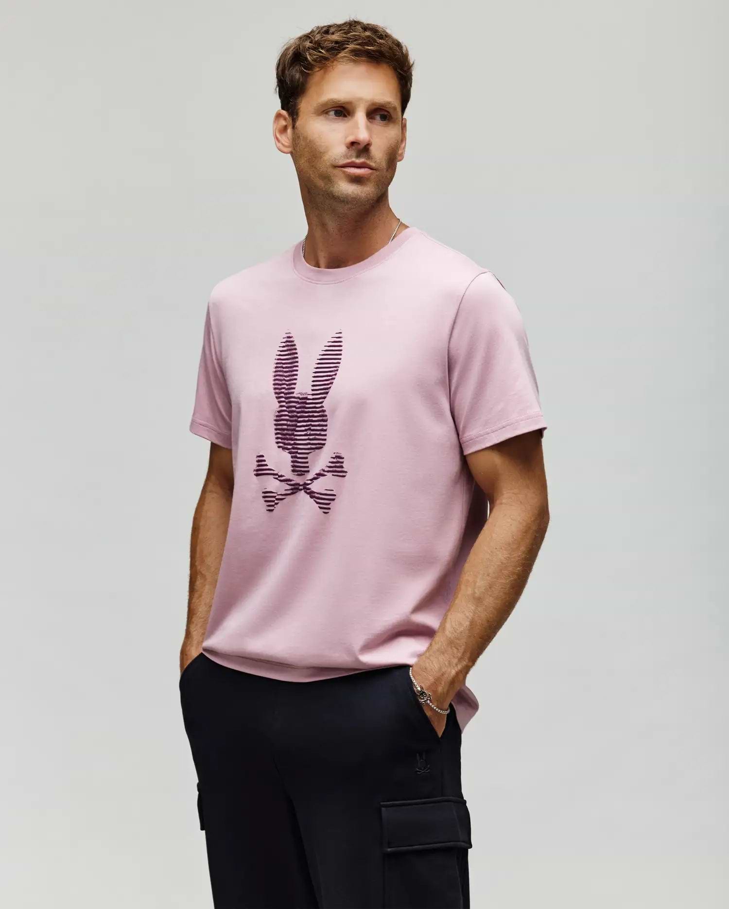 A man wears the Psycho Bunny MENS GRANT BUNNY GRAPHIC TEE (MT0200081), a light pink Pima cotton shirt with a stylized bunny and crossbones, paired with black pants, standing hands-in-pockets against a plain light background. (2)