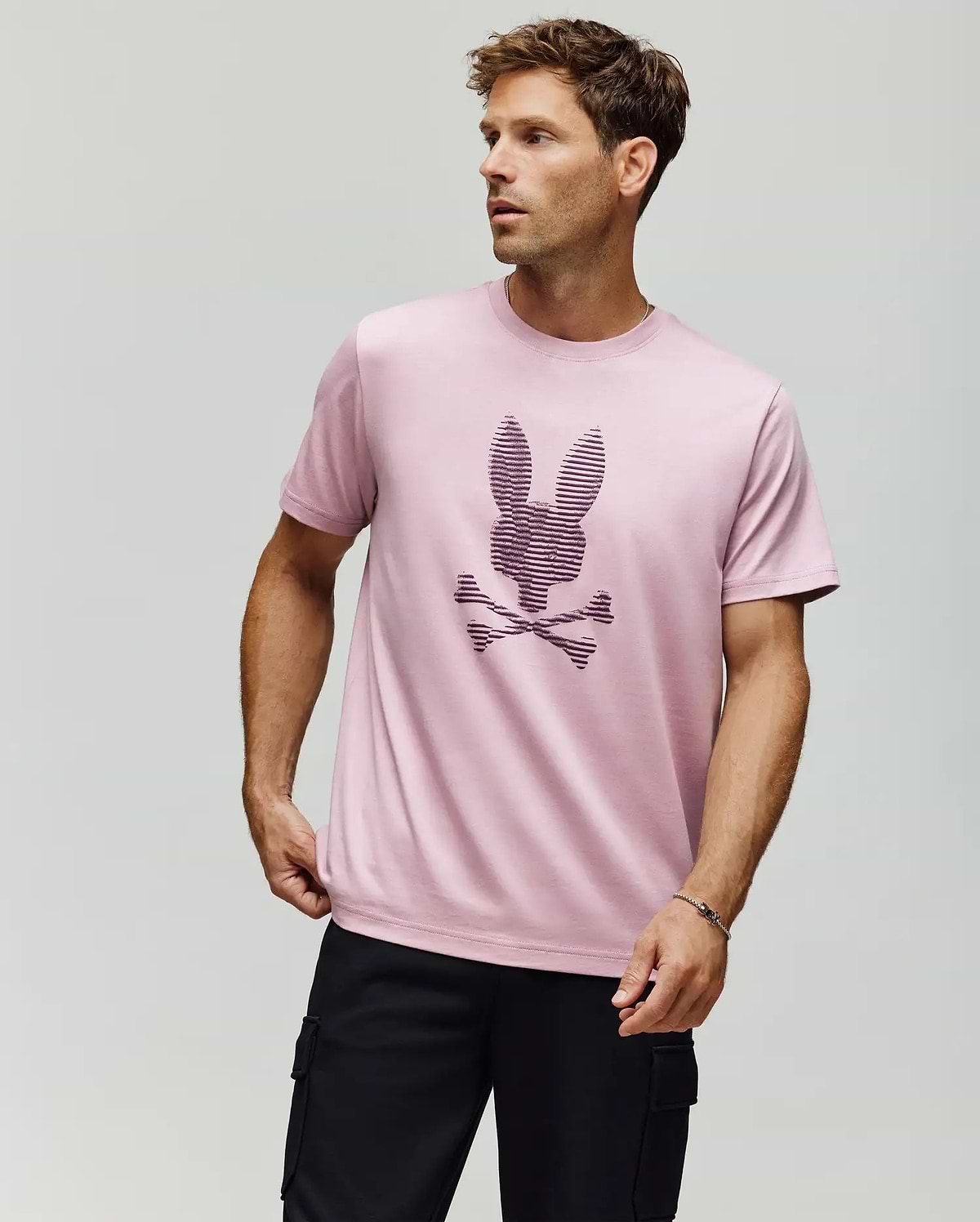 A man stands against a plain background wearing the Psycho Bunny MENS GRANT BUNNY GRAPHIC TEE (MT0200081) in light pink, made from Pima cotton with a textured bunny head above crossed bones, paired with black cargo pants. (4)