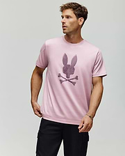 A man stands against a plain background wearing the Psycho Bunny MENS GRANT BUNNY GRAPHIC TEE (MT0200081) in light pink, made from Pima cotton with a textured bunny head above crossed bones, paired with black cargo pants. (4)