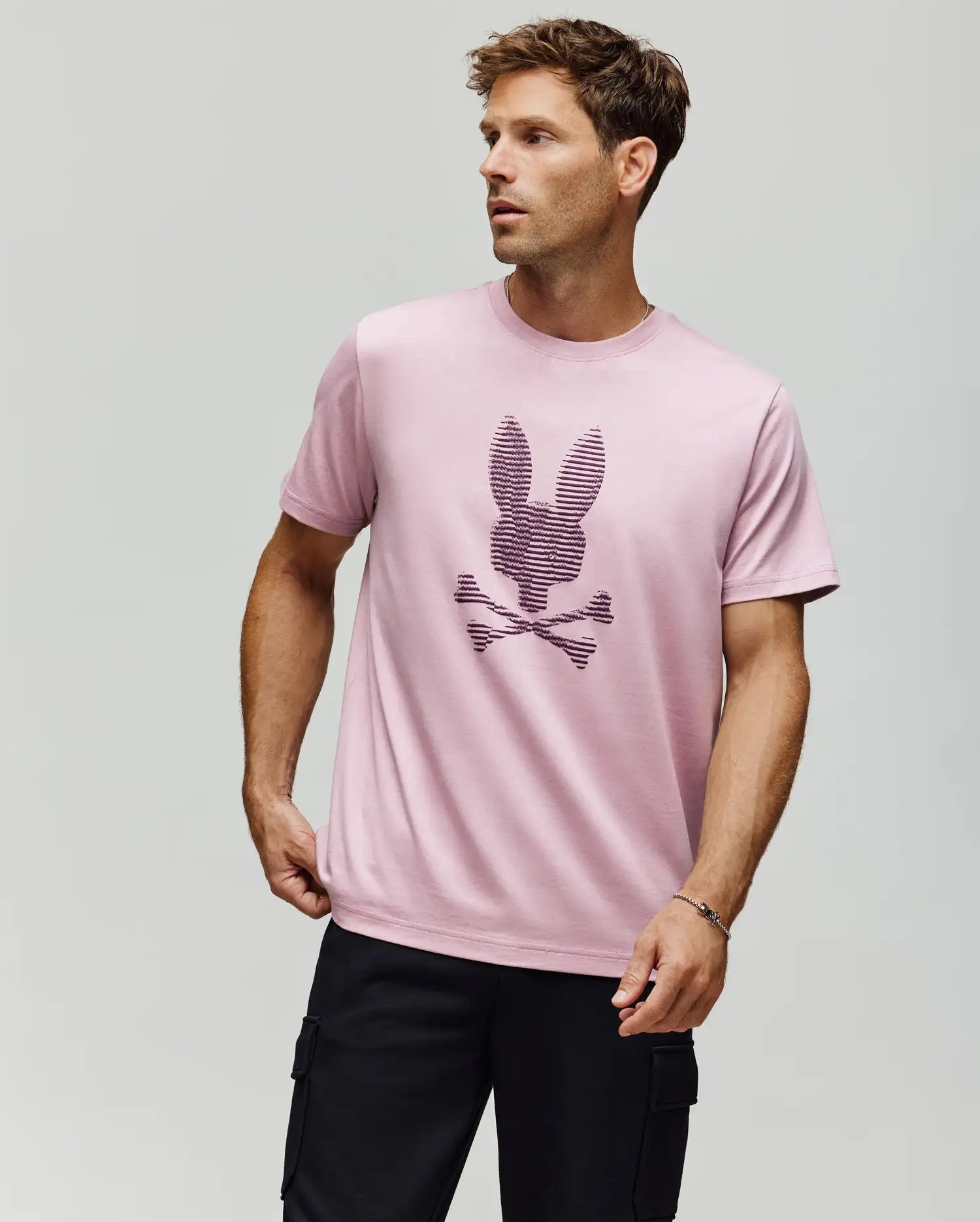 A man stands against a plain background wearing the Psycho Bunny MENS GRANT BUNNY GRAPHIC TEE (MT0200081) in light pink, made from Pima cotton with a textured bunny head above crossed bones, paired with black cargo pants. (4)
