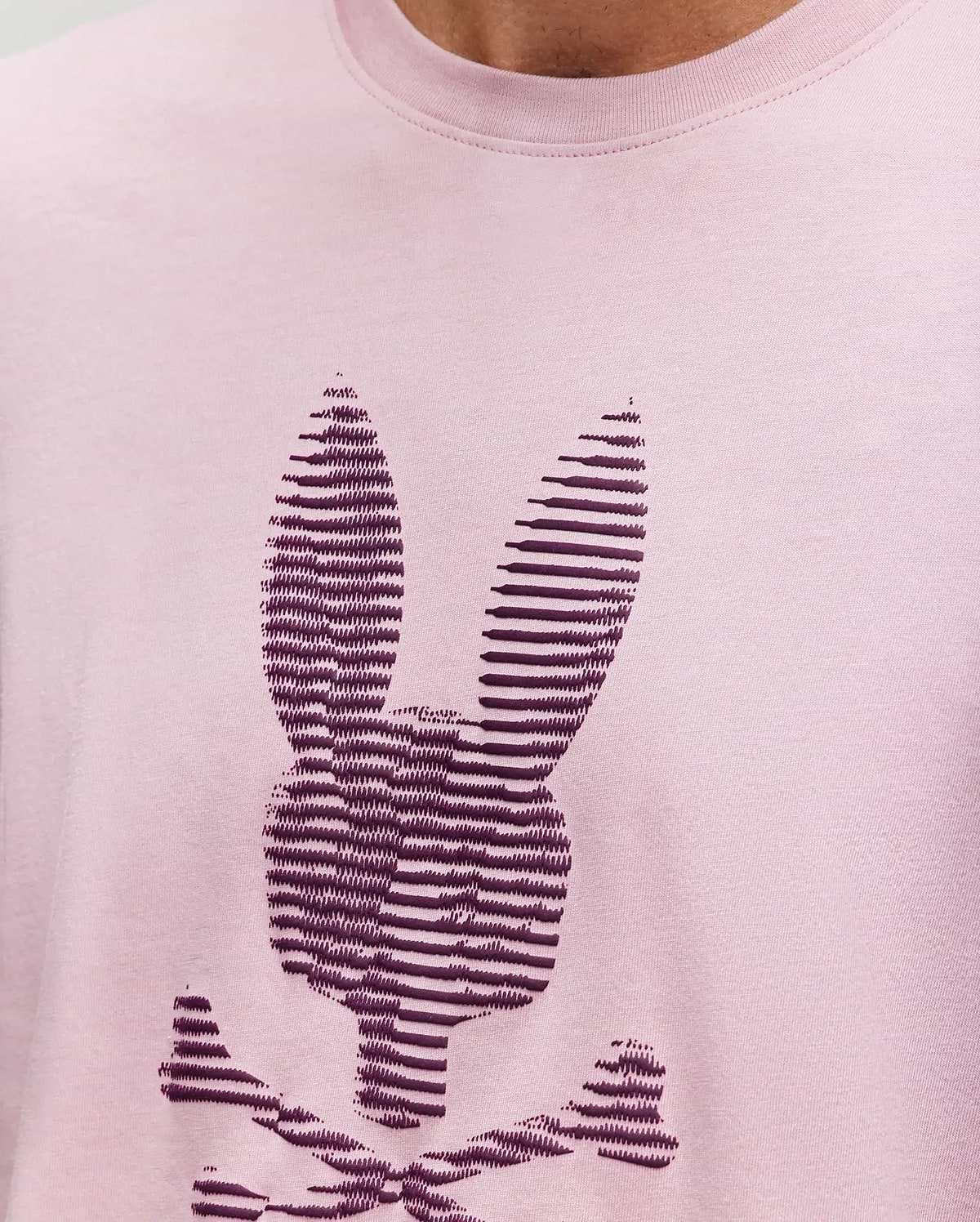 A close-up of the Psycho Bunny MENS GRANT BUNNY GRAPHIC TEE, crafted in Peru from soft Pima cotton, shows a stylized dark purple bunny head with crossbones in horizontal stripes on light pink fabric. (3)
