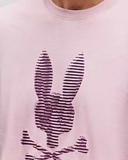 A close-up of the Psycho Bunny MENS GRANT BUNNY GRAPHIC TEE, crafted in Peru from soft Pima cotton, shows a stylized dark purple bunny head with crossbones in horizontal stripes on light pink fabric. (3)