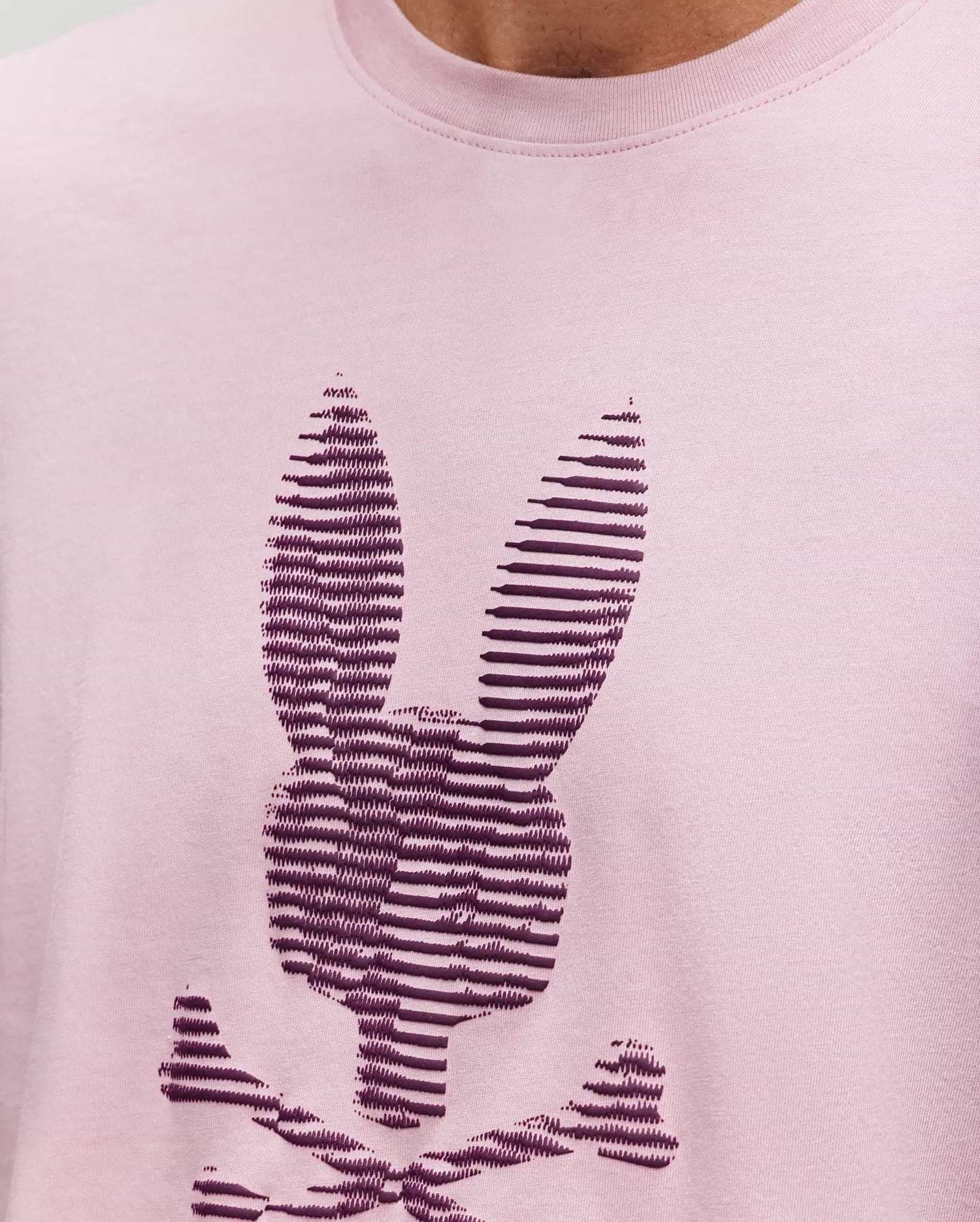 A close-up of the Psycho Bunny MENS GRANT BUNNY GRAPHIC TEE, crafted in Peru from soft Pima cotton, shows a stylized dark purple bunny head with crossbones in horizontal stripes on light pink fabric. (3)