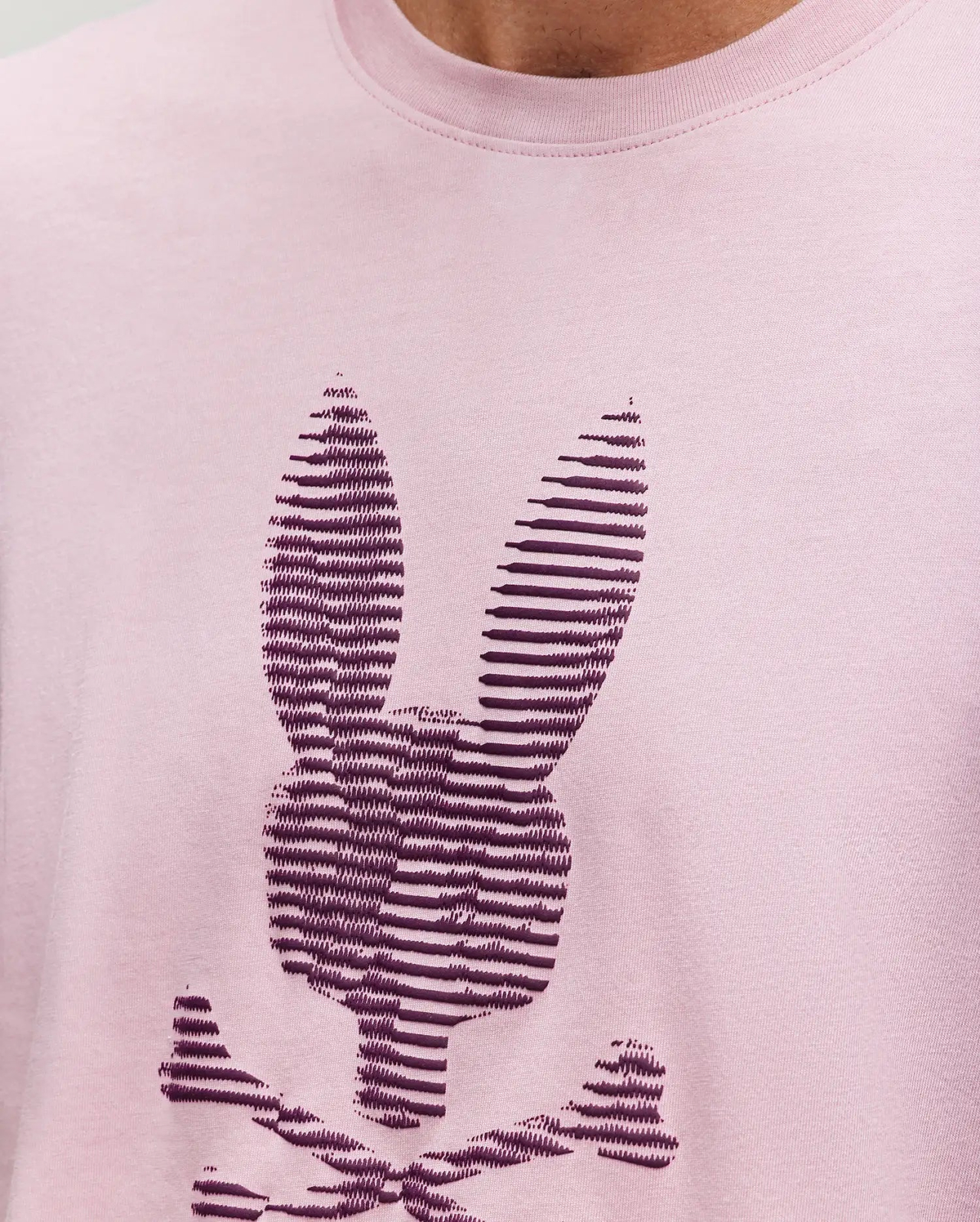 A close-up of the Psycho Bunny MENS GRANT BUNNY GRAPHIC TEE, crafted in Peru from soft Pima cotton, shows a stylized dark purple bunny head with crossbones in horizontal stripes on light pink fabric. (3)