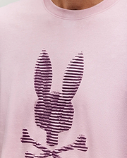 A close-up of the Psycho Bunny MENS GRANT BUNNY GRAPHIC TEE, crafted in Peru from soft Pima cotton, shows a stylized dark purple bunny head with crossbones in horizontal stripes on light pink fabric. (3)