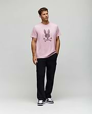 A man models the Psycho Bunny MENS GRANT BUNNY GRAPHIC TEE - MT0200081, made from soft Pima cotton in Peru, styled with black pants and black-and-white sneakers. He stands before a plain background with his left hand in his pocket, gazing to the side. (5)