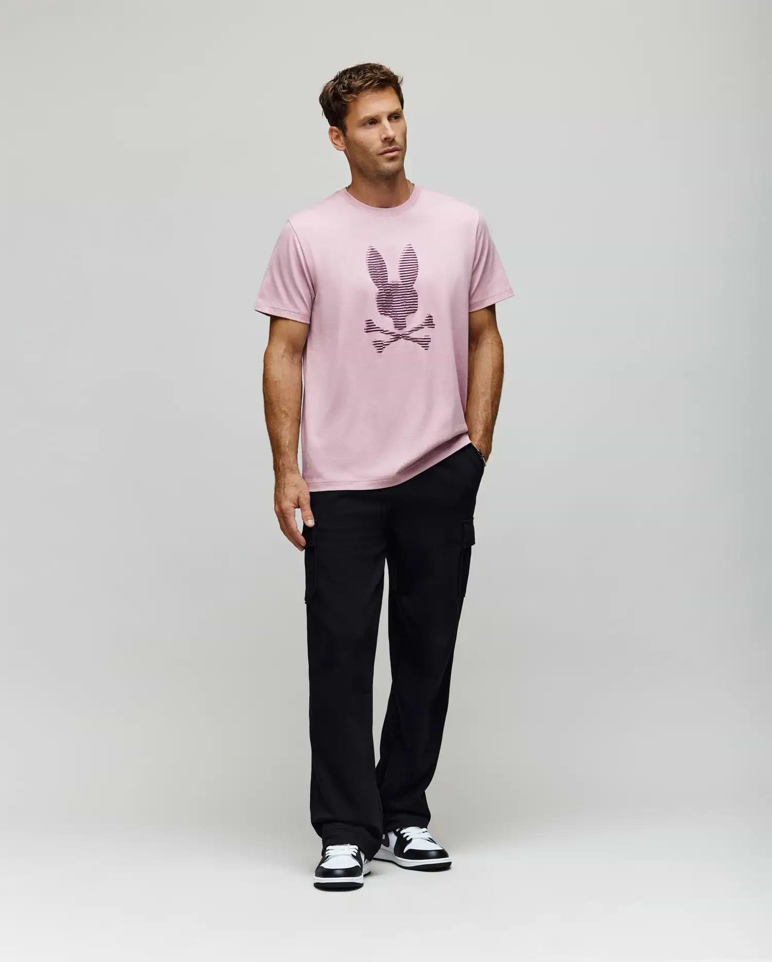 A man models the Psycho Bunny MENS GRANT BUNNY GRAPHIC TEE - MT0200081, made from soft Pima cotton in Peru, styled with black pants and black-and-white sneakers. He stands before a plain background with his left hand in his pocket, gazing to the side. (5)