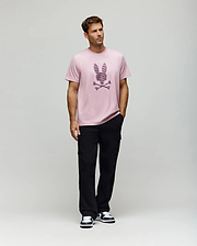 A man models the Psycho Bunny MENS GRANT BUNNY GRAPHIC TEE - MT0200081, made from soft Pima cotton in Peru, styled with black pants and black-and-white sneakers. He stands before a plain background with his left hand in his pocket, gazing to the side. (5)