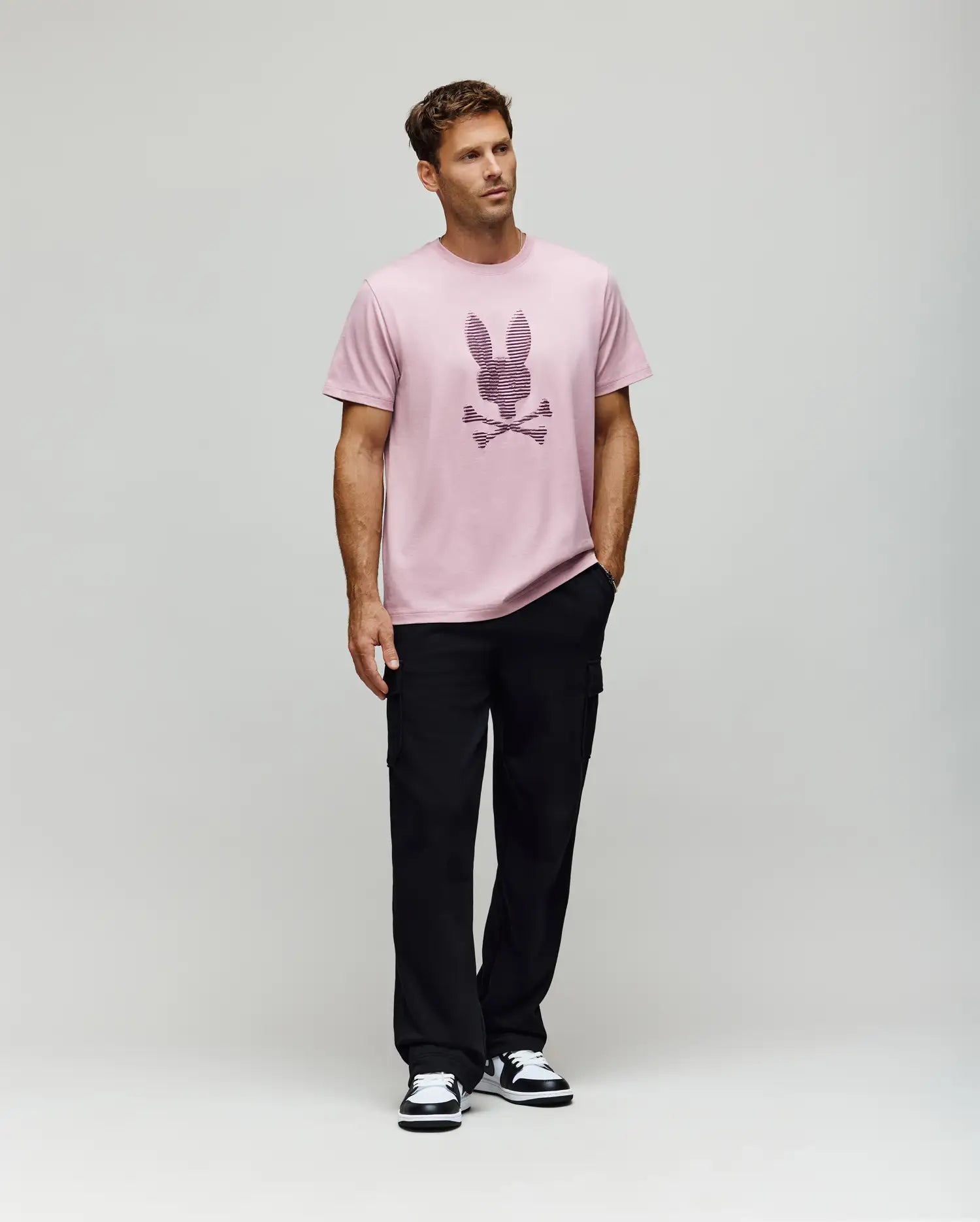 A man models the Psycho Bunny MENS GRANT BUNNY GRAPHIC TEE - MT0200081, made from soft Pima cotton in Peru, styled with black pants and black-and-white sneakers. He stands before a plain background with his left hand in his pocket, gazing to the side. (5)