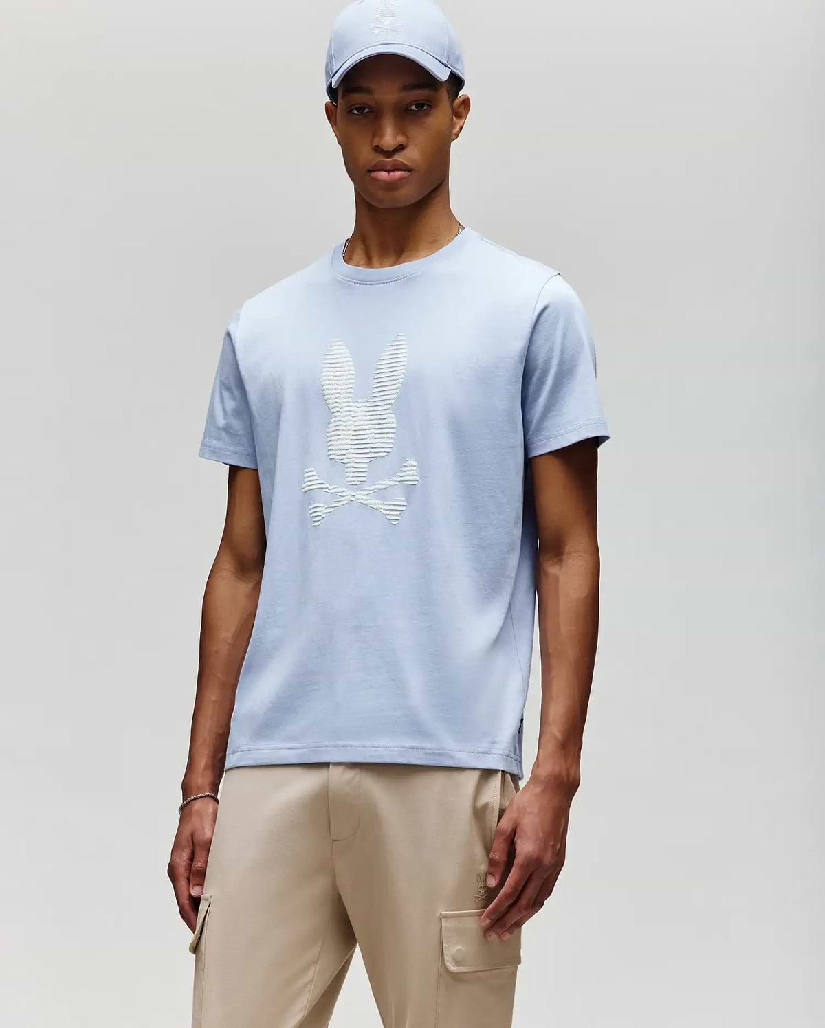 A man stands against a light gray background wearing a Psycho Bunny MENS GRANT BUNNY GRAPHIC TEE - MT0200081 in light blue with a matching cap, styled with beige cargo pants for a modern men's fashion look. (2)