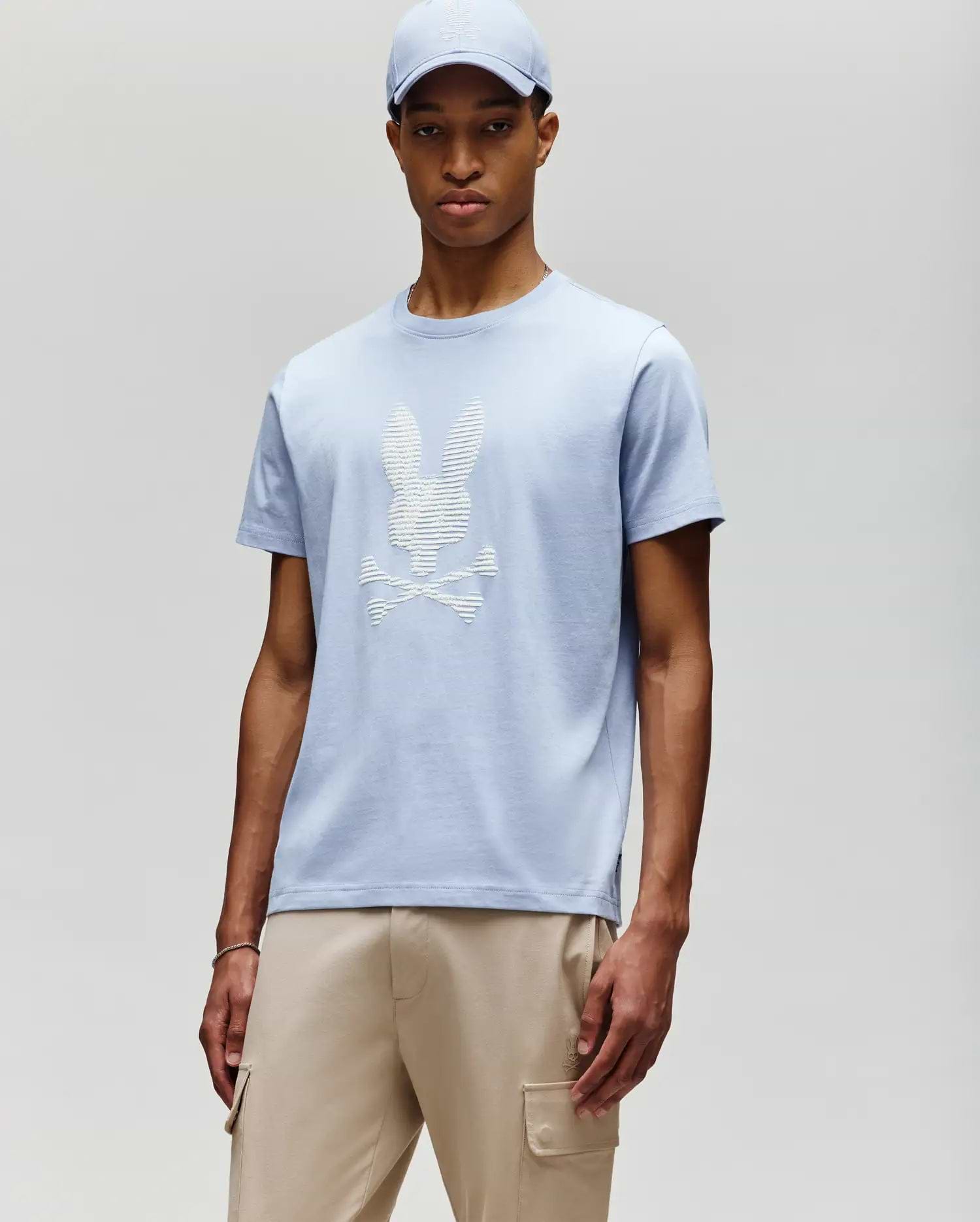 A man stands against a light gray background wearing a Psycho Bunny MENS GRANT BUNNY GRAPHIC TEE - MT0200081 in light blue with a matching cap, styled with beige cargo pants for a modern men's fashion look. (2)