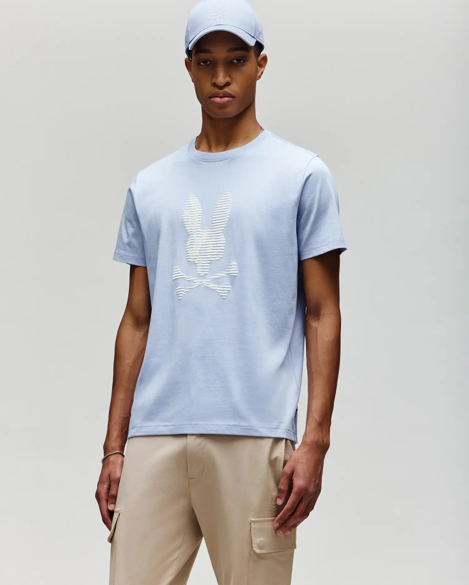 A man stands against a light gray background wearing a Psycho Bunny MENS GRANT BUNNY GRAPHIC TEE - MT0200081 in light blue with a matching cap, styled with beige cargo pants for a modern men's fashion look. (2)