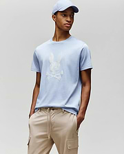 A man models Psycho Bunny’s MENS GRANT BUNNY GRAPHIC TEE - MT0200081 in light blue with a matching cap and beige drawstring pants, hands in pockets against a plain backdrop—an easy, stylish men's fashion look. (4)