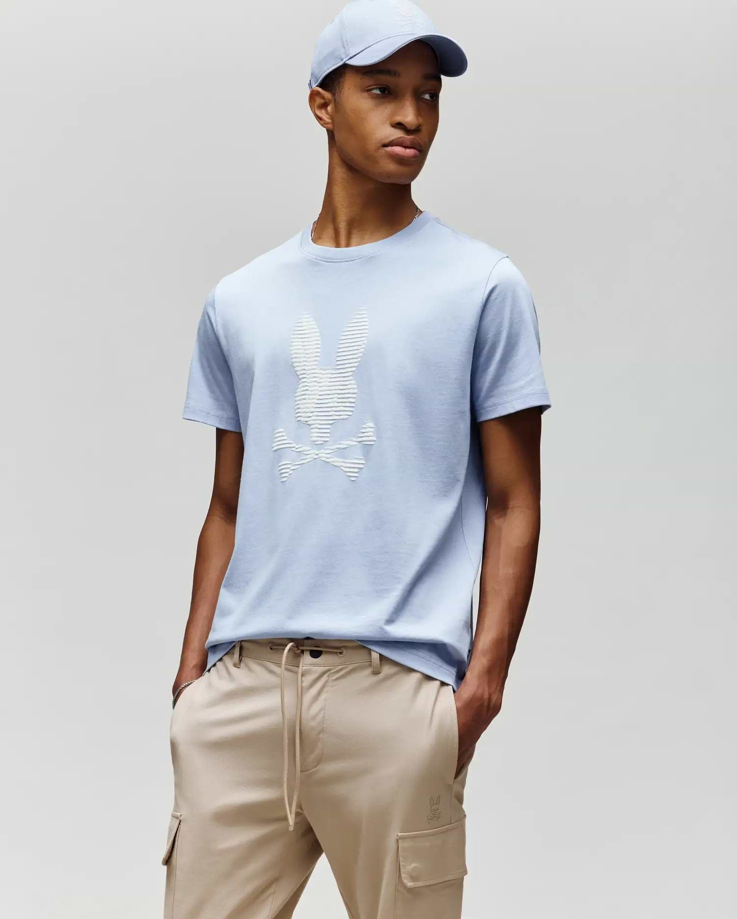 A man models Psycho Bunny’s MENS GRANT BUNNY GRAPHIC TEE - MT0200081 in light blue with a matching cap and beige drawstring pants, hands in pockets against a plain backdrop—an easy, stylish men's fashion look. (4)