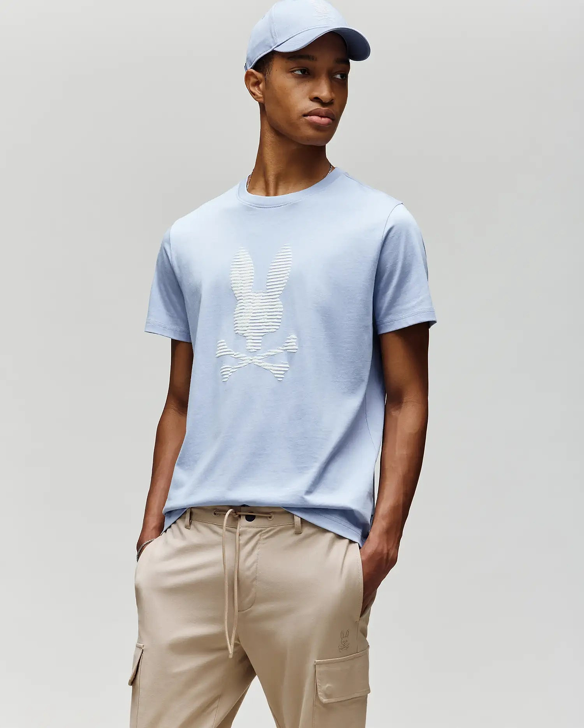 A man models Psycho Bunny’s MENS GRANT BUNNY GRAPHIC TEE - MT0200081 in light blue with a matching cap and beige drawstring pants, hands in pockets against a plain backdrop—an easy, stylish men's fashion look. (4)
