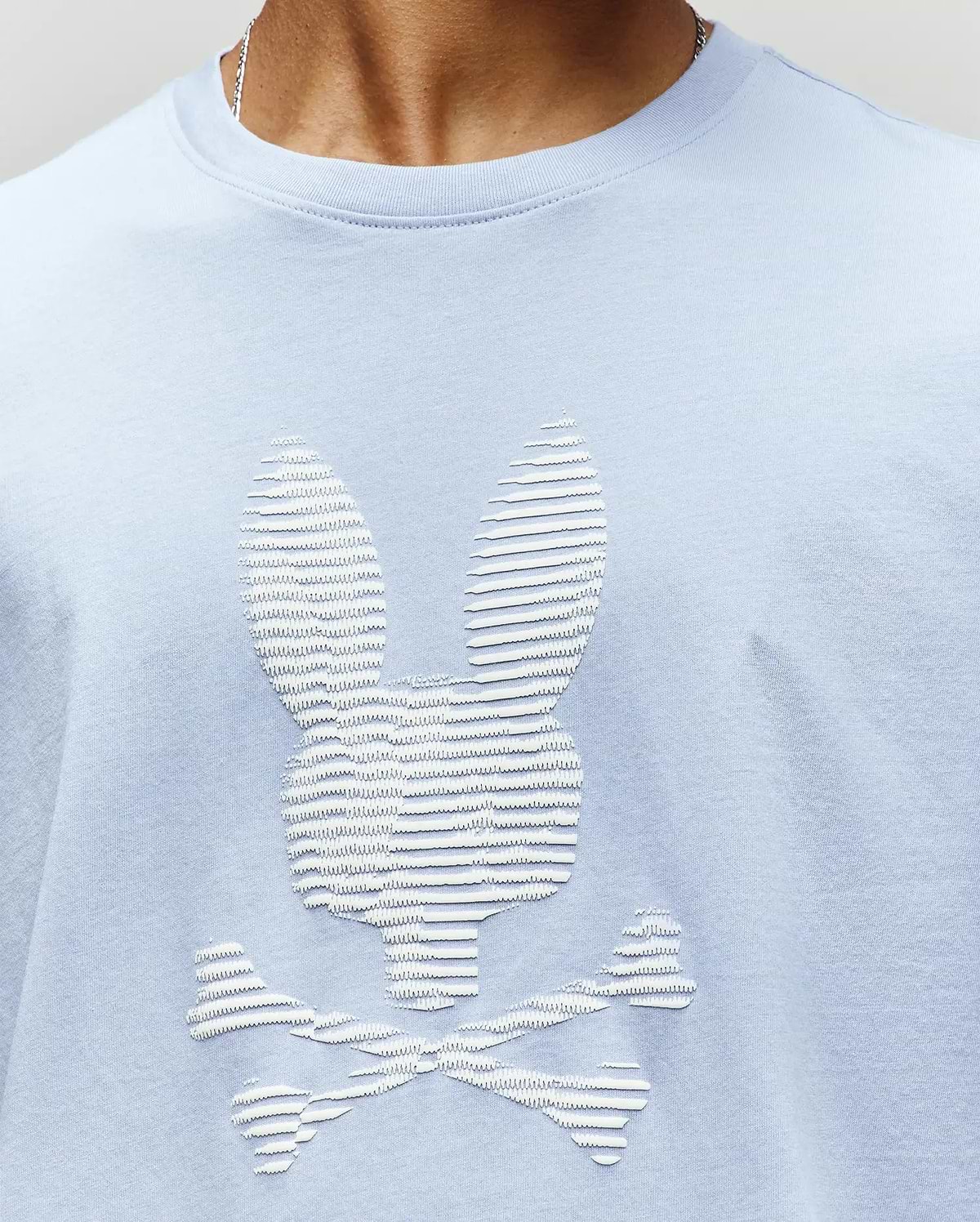 A man wears the Psycho Bunny MENS GRANT BUNNY GRAPHIC TEE (MT0200081), a light blue Pima cotton shirt featuring a white textured bunny head above crossed bones with raised horizontal lines—a bold choice in men’s fashion. (3)