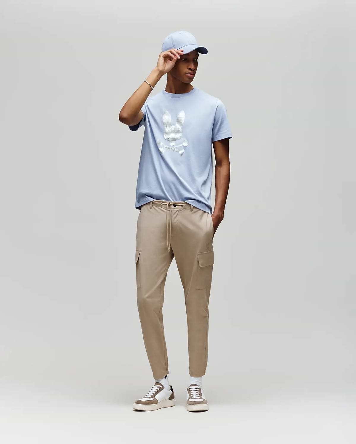 A man models the Psycho Bunny MENS GRANT BUNNY GRAPHIC TEE (MT0200081) in light blue, paired with beige cargo joggers and white sneakers, posed against a plain light gray background for a casual men's fashion look. (5)