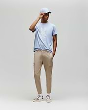 A man models the Psycho Bunny MENS GRANT BUNNY GRAPHIC TEE (MT0200081) in light blue, paired with beige cargo joggers and white sneakers, posed against a plain light gray background for a casual men's fashion look. (5)