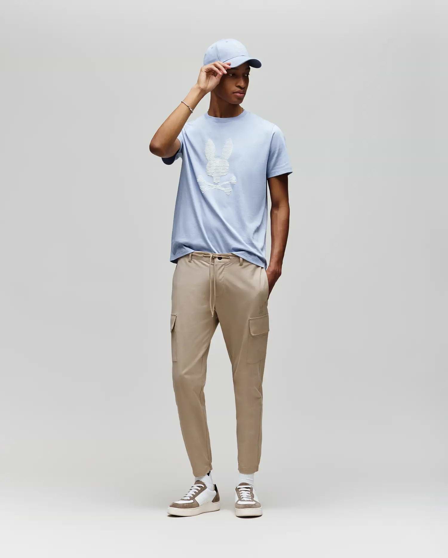 A man models the Psycho Bunny MENS GRANT BUNNY GRAPHIC TEE (MT0200081) in light blue, paired with beige cargo joggers and white sneakers, posed against a plain light gray background for a casual men's fashion look. (5)