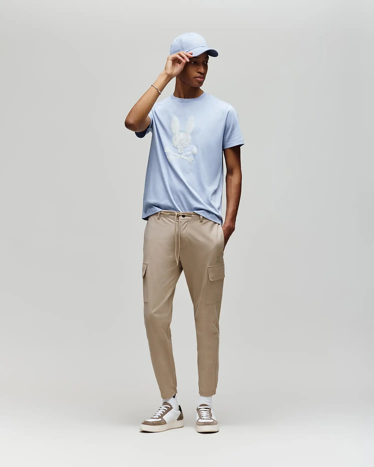 A man models the Psycho Bunny MENS GRANT BUNNY GRAPHIC TEE (MT0200081) in light blue, paired with beige cargo joggers and white sneakers, posed against a plain light gray background for a casual men's fashion look. (5)