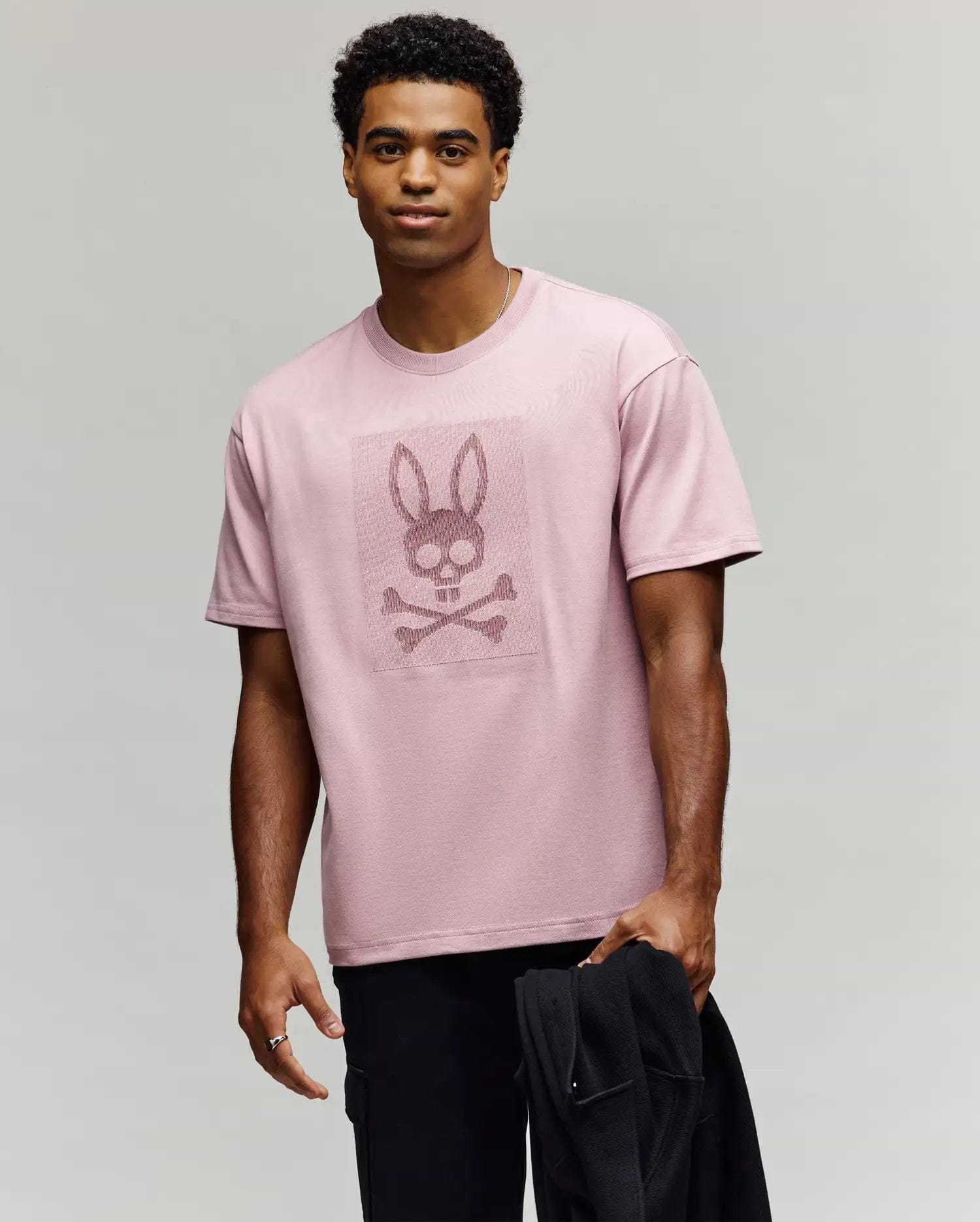 A man wears the Psycho Bunny MENS REECE OVERSIZED TEE (MT0200082), a light pink, relaxed-fit Pima cotton shirt with a stylized bunny skull and crossbones graphic. He holds a black jacket and looks at the camera, smiling slightly. (1)