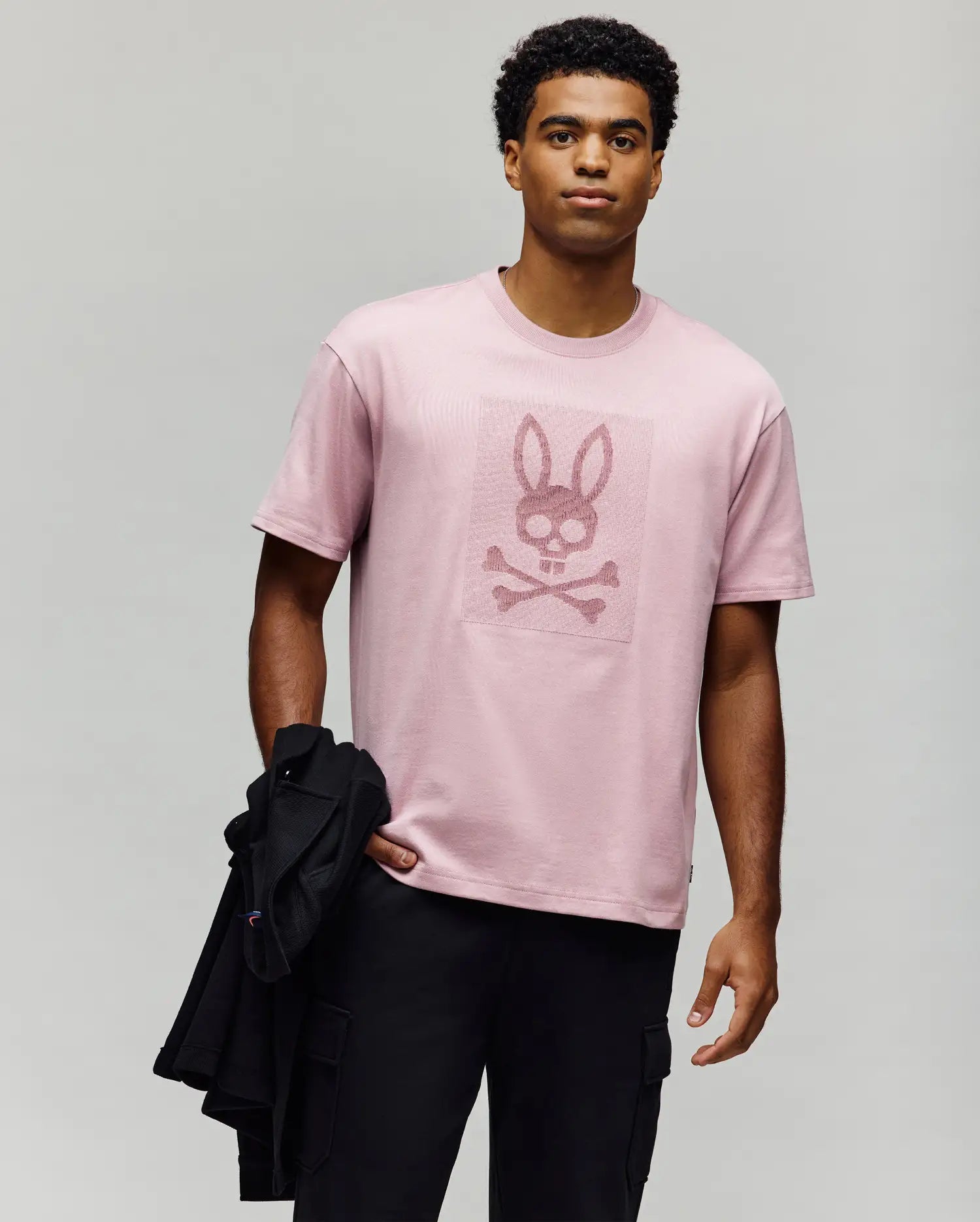 A man stands against a plain background wearing the Psycho Bunny MENS REECE OVERSIZED TEE (MT0200082), a relaxed fit, heavyweight Pima cotton tee with a bold textured bunny skull and crossbones graphic, paired with black pants and holding a black jacket. (3)