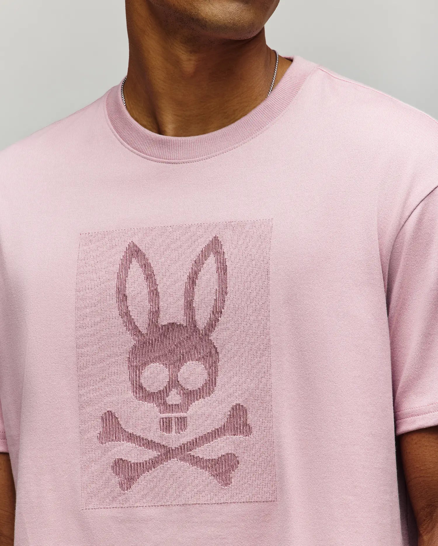 A person wears the Psycho Bunny MENS REECE OVERSIZED TEE (MT0200082), a light pink heavyweight jersey with a relaxed fit, featuring an embossed bunny skull and crossbones on the chest; their face is partially out of frame. (2)