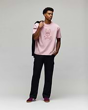 A man poses against a gray background wearing Psycho Bunny’s MENS REECE OVERSIZED TEE - MT0200082 in light pink Pima cotton with a bunny skull graphic, paired with black cargo pants, maroon sneakers, and holding a jacket over his shoulder. (4)