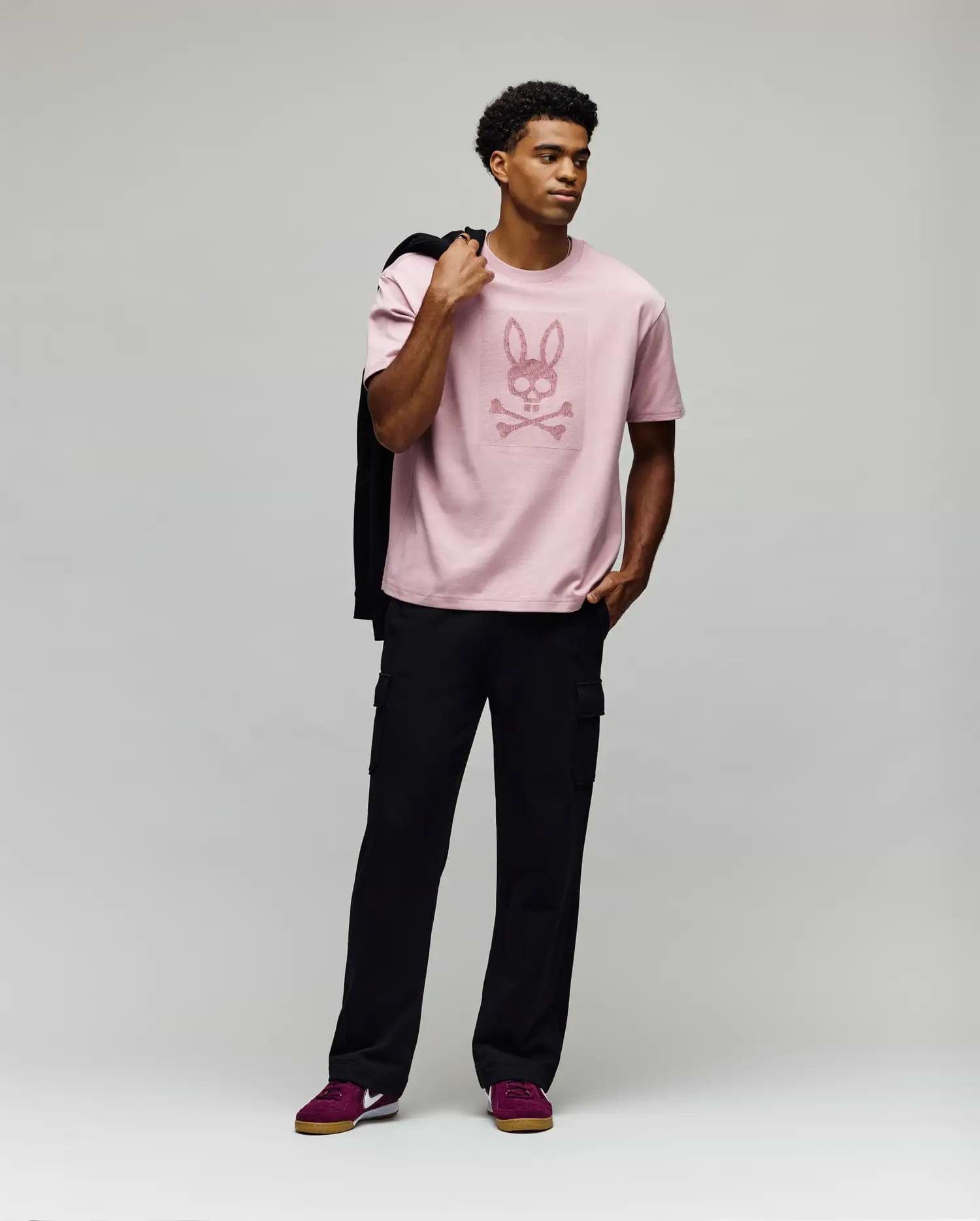 A man poses against a gray background wearing Psycho Bunny’s MENS REECE OVERSIZED TEE - MT0200082 in light pink Pima cotton with a bunny skull graphic, paired with black cargo pants, maroon sneakers, and holding a jacket over his shoulder. (4)