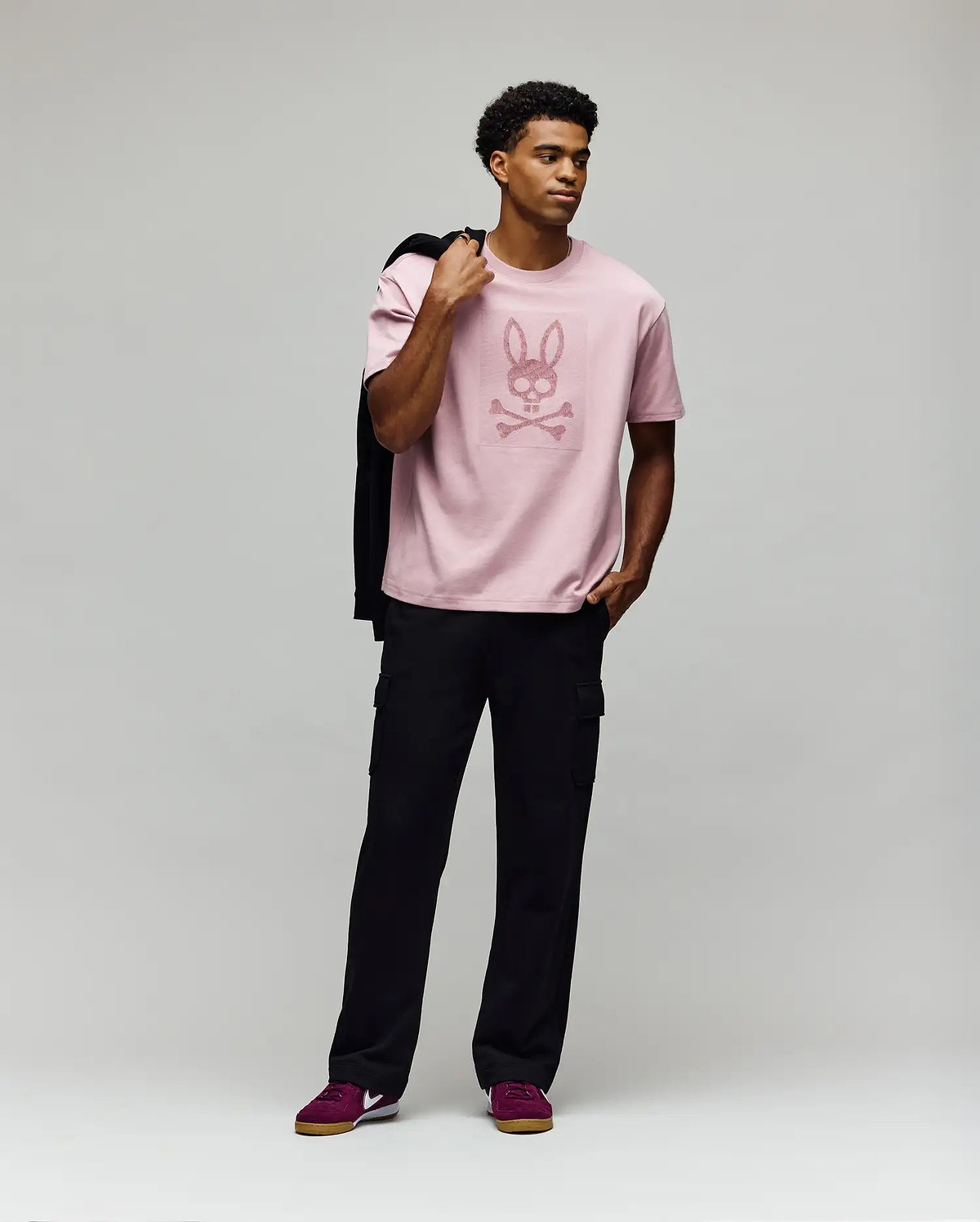 A man poses against a gray background wearing Psycho Bunny’s MENS REECE OVERSIZED TEE - MT0200082 in light pink Pima cotton with a bunny skull graphic, paired with black cargo pants, maroon sneakers, and holding a jacket over his shoulder. (4)