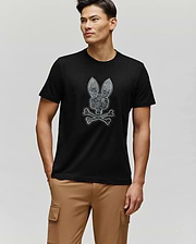 A man wearing the Psycho Bunny MENS ROWEN BUNNY GRAPHIC TEE (MT0200101), made of soft Pima cotton and featuring a bunny skull and crossbones graphic, stands with his right hand in beige cargo pants, looking left against a plain light background.