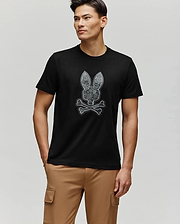 A man wearing the Psycho Bunny MENS ROWEN BUNNY GRAPHIC TEE (MT0200101), made of soft Pima cotton and featuring a bunny skull and crossbones graphic, stands with his right hand in beige cargo pants, looking left against a plain light background.