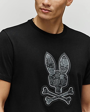 A person wears the Psycho Bunny MENS ROWEN BUNNY GRAPHIC TEE - MT0200101, a black Pima cotton tee featuring a bold floral Bunny patch with crossed bones on the chest. The person's face is partially visible.