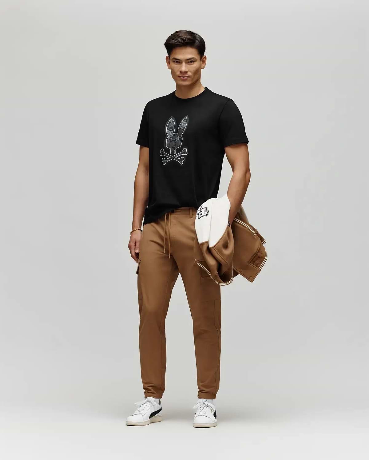 A man stands against a plain background wearing a black Psycho Bunny MENS ROWEN BUNNY GRAPHIC TEE (MT0200101) with a floral Bunny patch, brown joggers, white sneakers, and holding a brown and white jacket.