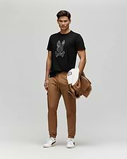 A man stands against a plain background wearing a black Psycho Bunny MENS ROWEN BUNNY GRAPHIC TEE (MT0200101) with a floral Bunny patch, brown joggers, white sneakers, and holding a brown and white jacket.