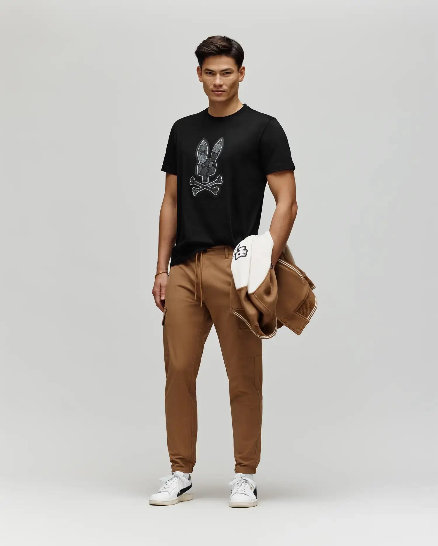A man stands against a plain background wearing a black Psycho Bunny MENS ROWEN BUNNY GRAPHIC TEE (MT0200101) with a floral Bunny patch, brown joggers, white sneakers, and holding a brown and white jacket.
