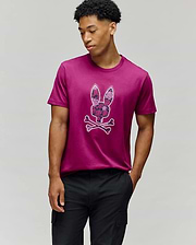 A man stands against a plain gray background wearing the Psycho Bunny MENS ROWEN BUNNY GRAPHIC TEE (MT0200101), a magenta Pima cotton shirt with a bunny skull and crossbones graphic, one hand in his pocket as he looks down to the side. (2)